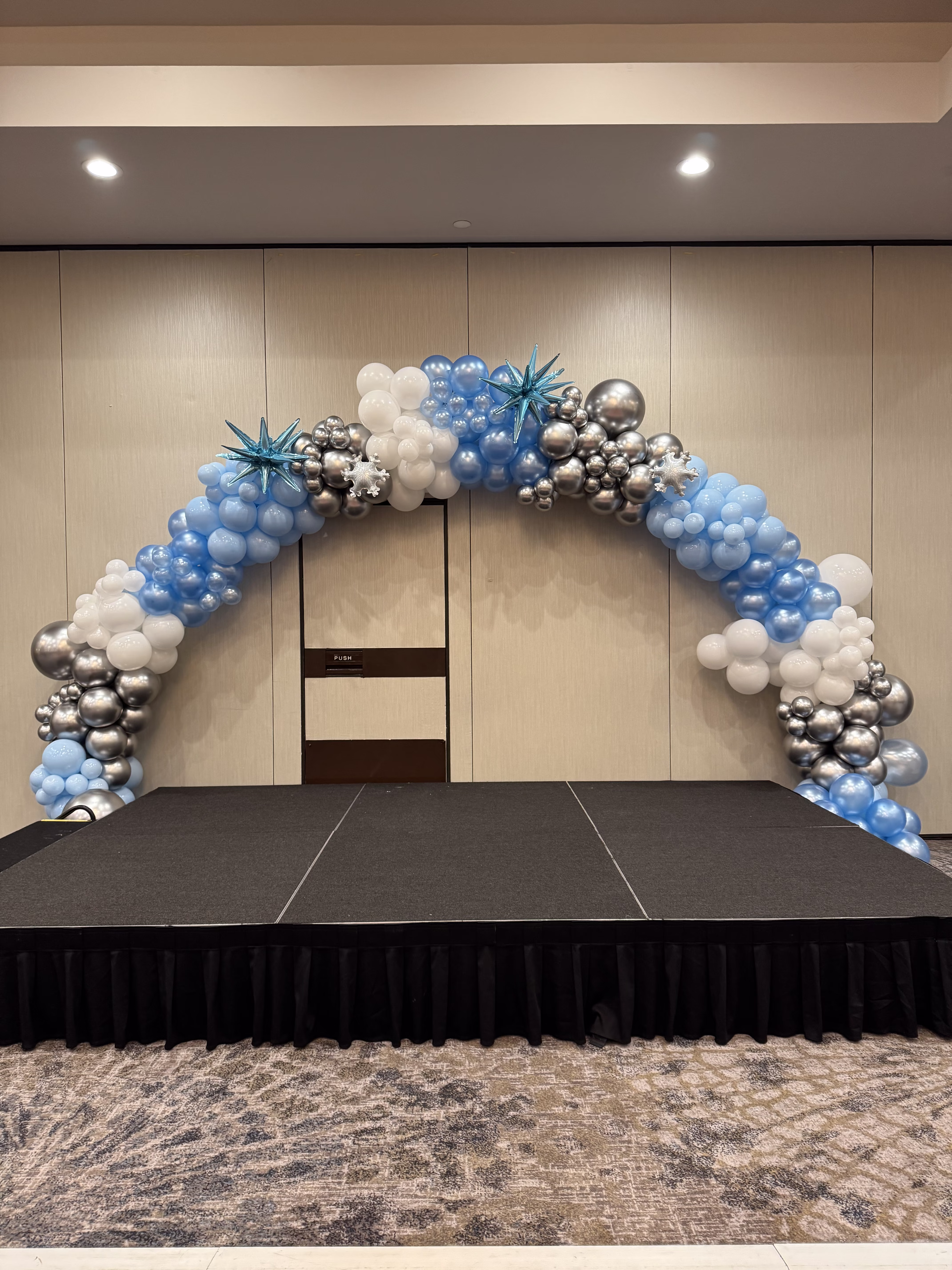 Arco de globos azul, blanco y plateado sobre escenario en salón de hotel para evento corporativo